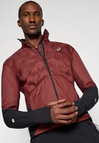 Giacca isolante rossa e nera con colletto alto, texture trapuntata sulla parte superiore, maniche lunghe nere e chiusura con zip. Accenti riflettenti presenti.