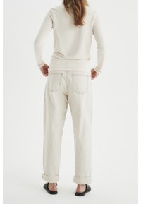 Kvinna står med händerna på höfterna iförd en beige långärmad topp, ljusa uppvikta byxor och svarta slip-on skor, med ryggen mot kameran.