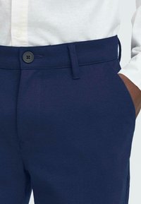 Jūras zila teksturēta bikses ar pogas aizdari jostasvietā. Jostas vietā ir jostas cilpas, un roka ir novietota sānu kabatā.