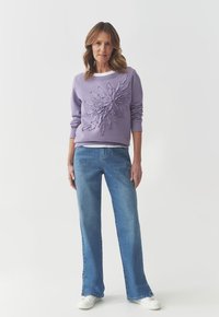 Felpa lilla con design floreale testurizzato, collo a giro e maniche lunghe. Abbinata a jeans svasati di un blu chiaro e sneakers bianche.