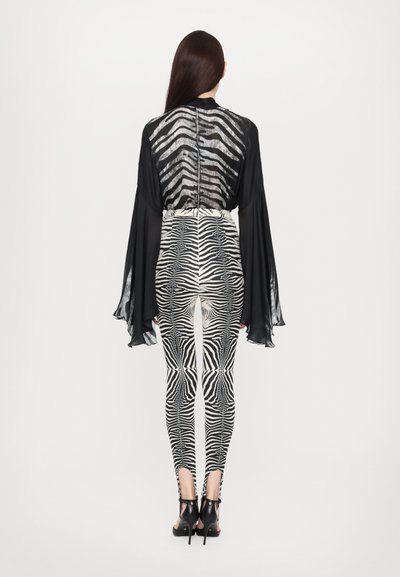 Bluză neagră transparentă cu mâneci lungi și spate cu dungi, asortată cu colanți cu imprimeu zebra alb-negru; pantofii cu toc negri completează look-ul.
