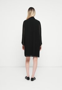 Μαύρη φούστα φόρ�εμα με μακριά μανίκια. Ελαφρύ, διάφανο ύφασμα που δημιουργεί κάθετες γραμμές. Σημαντική λεπτομέρεια: ελαφρώς καμπυλωτό τελείωμα. Φορεμένο με μαύρες παπούτσια.