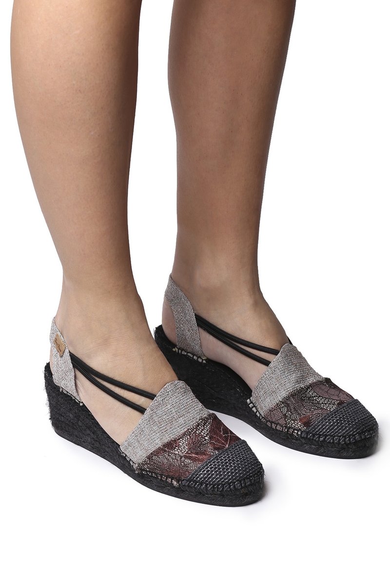 Jambes portant des espadrilles compensées en tissu de dentelle beige, noir et bordeaux, avec des sangles élastiques noires et des semelles tissées noires.