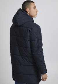 Jūras spēku pufotais uzvalks ar kapuci, ar horizontāliem quilted līmeņiem, pilna garuma rāvējslēdzis un sānu atveres papildu komfortam.