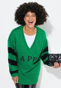 Pullover verde lavorato a maglia con scollatura a V, strisce nere sulle maniche e la scritta "HAPPY" stampata sul davanti. Modella che tiene una borsa a tracolla nera.