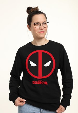 Kobieta w okularach i z kokiem na włosach, ubrana w czarną bluzę Deadpool, trzymająca za dół, na jednolitym jasnym tle.