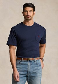 Mann iført marineblå kortermet t-skjorte med brystlomme og rødt logo, lyseblå jeans, brunt belte og armbåndsur, stående med hånden i lommen.