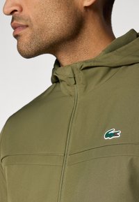Sweat-shirt zippé de couleur olive avec une texture, arborant un petit logo crocodile vert et rouge sur la poitrine gauche.