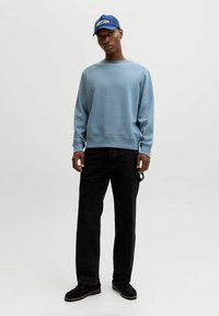 Sudadera azul claro con cuello redondo y puños acanalados, combinada con vaqueros negros y zapatos negros. La modelo lleva una gorra azul con texto blanco.