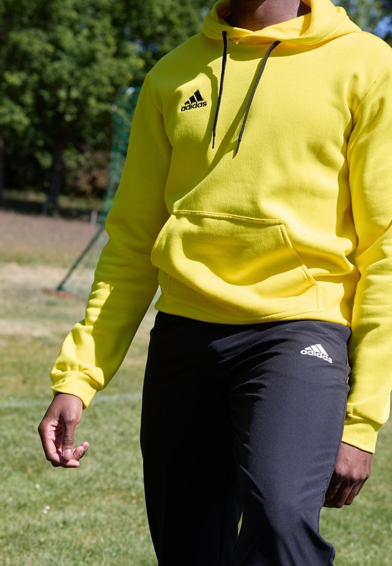 Personne portant un sweat à capuche jaune Adidas et un pantalon noir Adidas, debout sur l'herbe avec des arbres et un but de football en arrière-plan.