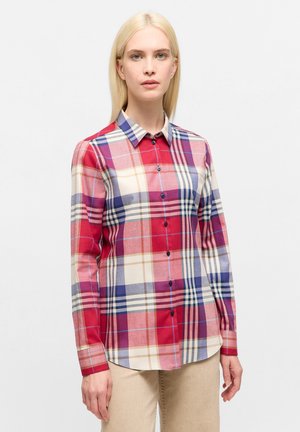 Donna bionda che indossa una camicia button-up a maniche lunghe con un motivo a quadri rosso, viola, crema e beige, abbinata a pantaloni beige chiaro.