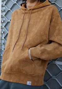 Persona con una sudadera marrón de Carhartt con patrón, con las manos en el bolsillo delantero, contra un fondo de rejilla metálica gris.