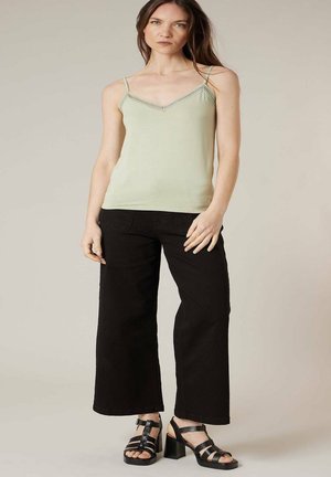 Femme debout portant un haut sans manches vert clair, un pantalon noir à jambes larges et des sandales à talons noires, devant un fond uni.