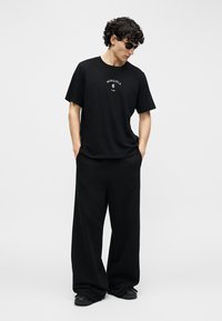 Mladý muž v čiernych slnečných okuliaroch, čiernom voľnom tričku s bielym nápisom, voľných čiernych nohaviciach, stojaci s rukami v vreckách.