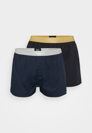 Dva páry boxerky. Jedny námořnické s stříbrným pruhem, jedny černé se zlatým pruhem. Obě mají hladký povrch a přední knoflík.