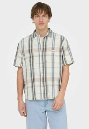 Jeune homme portant une chemise à manches courtes beige, bleue et grise à rayures verticales avec un jean bleu clair, debout devant un fond uni.