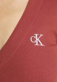 Karbowany top z głębokim dekoltem w kolorze rdzy z białym haftowanym logo "CK". Posiada dopasowany krój i miękką teksturę.