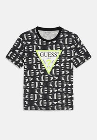 T-shirt noir en coton avec un motif répété et un grand triangle vert portant le logo "GUESS" en blanc, manches courtes, encolure ronde.