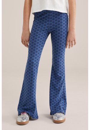 Laps, kes kannab siniste mustriliste laiade pükstega ning hõbedaste tossudega, seisab neutraalse taustaga.