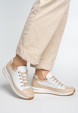 Feet wearing beige and white low-top sneakers with laces and side zippers, paired with beige corduroy pants rolled at the ankles.