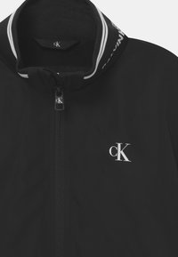 Černá bunda se zipem vpředu, vysokým límcem a logem. Má texturovanou tkaninu a bílé akcentní pruhy na límci.