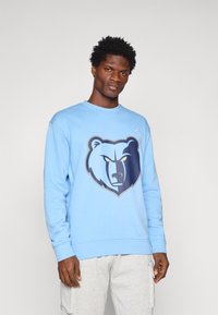 Világoskék pulóver, nagy, sötétkék és szürke medve grafikával. Pamut keverék, kerek nyakú dizájn, bordázott mandzsetta és szegély. Szürke nadrággal párosítva.