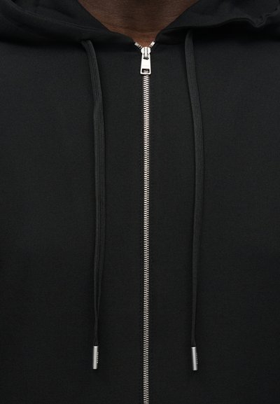 Μαύρο φούτερ με φερμουάρ και ασημένιο φερμουάρ, μαύρα κορδόνια με μεταλλικές άκρες, κοντινή λήψη στην περιοχή του στήθους και της κουκούλας.