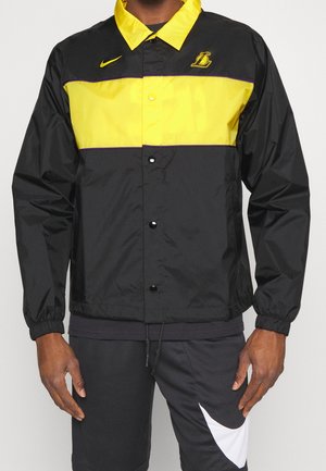 Mann trägt eine schwarze und gelbe Nike-Jacke mit dem Logo der Los Angeles Lakers, kombiniert mit schwarzen Shorts, die einen großen weißen Nike-Swoosh zeigen.