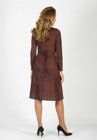 Brązowa sukienka z długim rękawem, z dopasowanym paskiem w talii, zdobnym ciemnym wzorem, spódnicą w kształcie litery A i długością do kolan. Nałożona z czarnymi szpilkami.