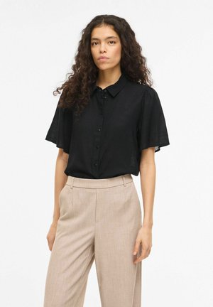 Kobieta o kręconych włosach ubrana w czarną, krótkorękawową, zapinaną na guziki bluzkę oraz beżowe, szerokie spodnie z wysokim stanem na jednolitym tle.