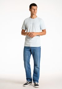 Helles blaues Baumwoll-T-Shirt mit kurzen Ärmeln, das den Markennamen in dunklerem Blau zeigt. Kombiniert mit geraden Jeans und schwarzen Sneakern.