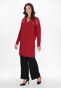 Femeie îmbrăcată cu un cardigan lung roșu cu butoni și umăr cu paiete, pantaloni negri cu picioare largi și tocuri negre, stând și zâmbind.