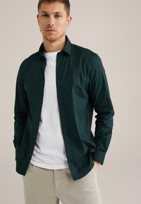 Donkergroen overhemd met lange mouwen en een button-down kraag, met een gladde textuur en zwarte knopen, in laagjes gedragen over een wit T-shirt.