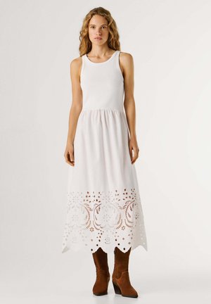 Vrouw met een mouwloze witte midi-jurk met een bloemenuitsnede aan de zoom en bruine suède laarzen tot halverwege de kuit, staand tegen een effen achtergrond.