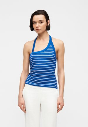 Kvinde iført blå og hvide stribede asymmetrisk haltertop med hvide bukser, stående mod en ensfarvet lys baggrund.