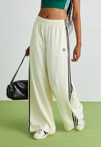 Calças largas cremes com riscas laterais pretas, cintura elástica e pequeno detalhe de logo. Usadas com uma bolsa preta e sapatos com detalhes prateados.
