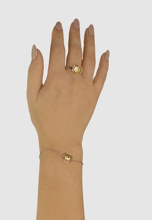 Gold-tone ring and bracelet set with circular designs featuring sunburst patterns, set on a slender silver chain and worn on a hand.