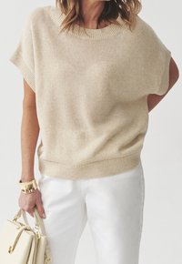 Donna che indossa un maglione a maglia beige a maniche corte, pantaloni bianchi, un bracciale d'oro e tiene una borsa crema su uno sfondo neutro.