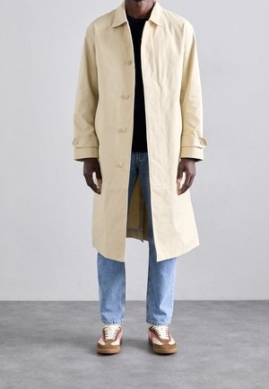 Uomo che indossa un lungo cappotto beige sopra una camicia nera, jeans azzurri e sneakers marroni e rosa, in piedi su pavimento grigio.