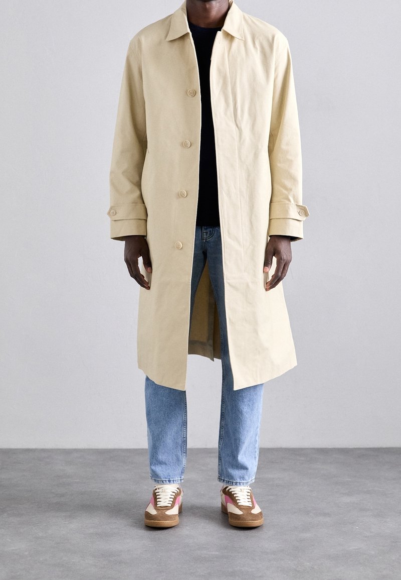 Homme portant un manteau long beige par-dessus une chemise noire, un jean bleu clair et des baskets marron et roses, debout sur un sol gris.