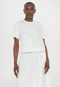 Top alb cropt cu mâneci scurte, având broderie florală. Materialul este neted cu un ușor luciu. Asortat cu o fustă albă corespunzătoare.