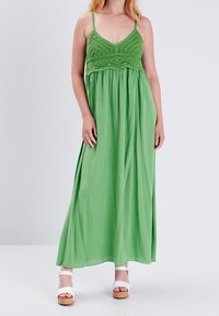 Femme portant une robe maxi verte sans manches avec un haut en crochet et des sandales plateformes blanches, debout devant un fond blanc uni.
