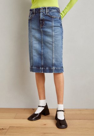 Kvinde iført en knælængde blå denim nederdel, hvide sokker, sorte Mary Jane sko og en limegrøn langærmet top med hænderne bag ryggen.