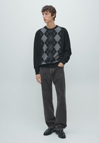 Schwarzer Strickpullover mit grauem Rautenmuster, kombiniert mit grauer Jeans und schwarzen Schuhen. Das Model steht mit den Händen in den Taschen.