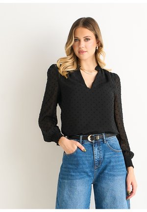 Black blouse with textured dot pattern, V-neckline, and puffed sleeves; paired with high-waisted blue denim jeans and a black belt.