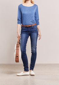 Vrouw in blauwe lange mouwen top, donkere blauwe jeans met een bruine gevlochten riem, witte instapschoenen, met een beige en bruine handtas aan de zijkant.
