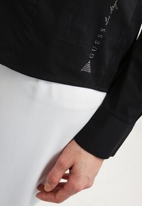 Czarna kurtka o gładkiej fakturze, z logo w srebrnych cyrkoniach i trójkątnym akcentem. Końce rękawów są lekko dopasowane.