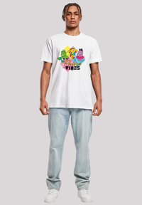 White cotton t-shirt with cartoon characters printed in vibrant colors. Design features "Positive Vibes" text and a geometric background. Worn with light blue jeans and white shoes.