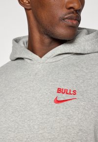 Grå hættetrøje med et rødt broderet "BULLS" logo og Nike swoosh. Stoffet er blødt og struktureret, med en hætte med snøre.