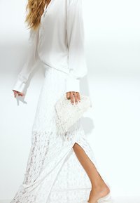 Hvid langærmet blouse parret med en hvid, blonde-mønsteret maxiskirt med en sideslids. Holder en tekstureret, frynset clutch.